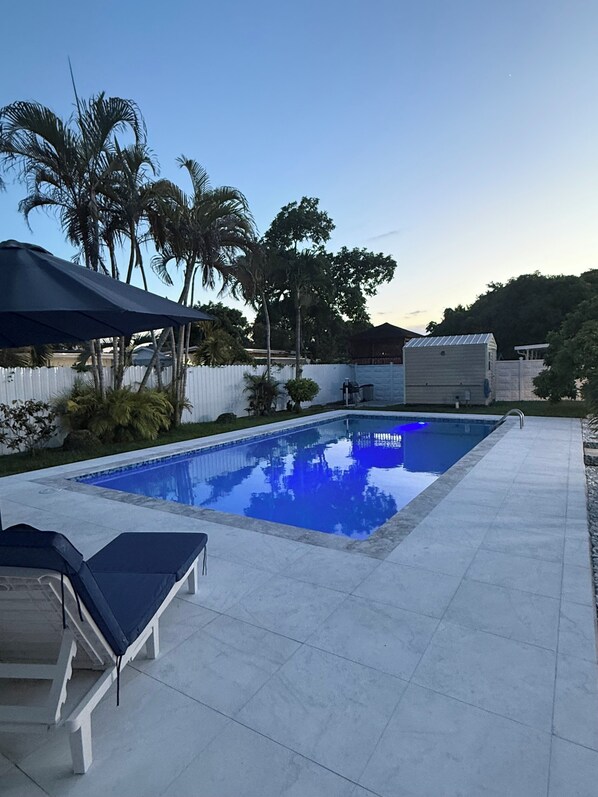Una piscina con plataforma de baldosas, una silla de descanso y una zona cubierta.