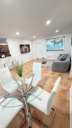 Una sala de estar moderna con una mesa de comedor de vidrio, sillas blancas, un sofá gris y un televisor montado en la pared.