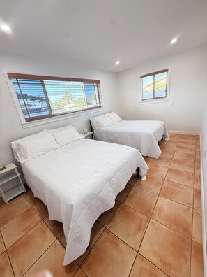 Una habitación con dos camas, una mesita de noche, una ventana con persianas y suelo de baldosas.