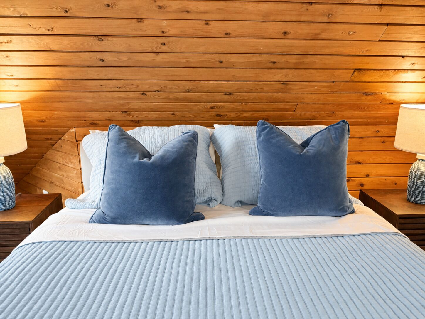 A bed with a blue quilt and pillows, wooden headboard, and two bedside lamps.