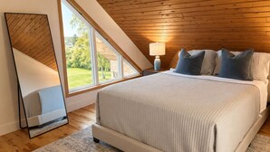 Quarto com cama grande, mesa de cabeceira, abajur e vista do exterior por um claraboia.