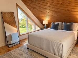 A bedroom with a large bed, a nightstand, a lamp, and a view of the outdoors through a skylight.