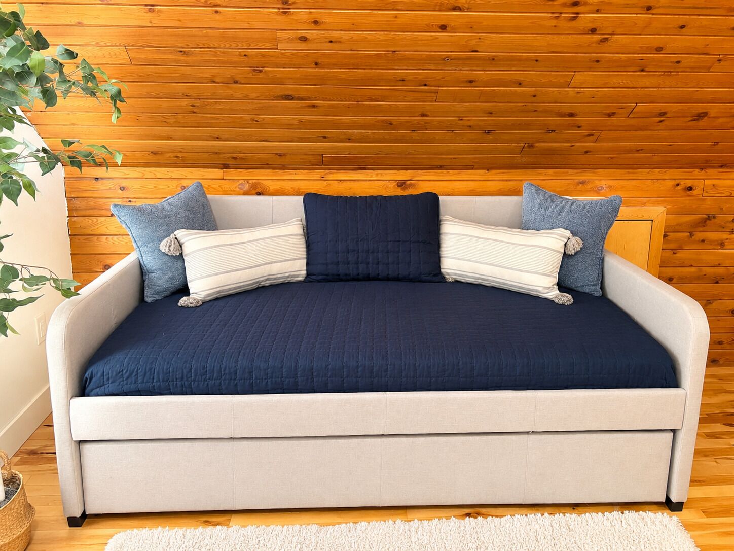 A sofa bed with a dark blue cover, light-colored cushions, and a wooden wall background.