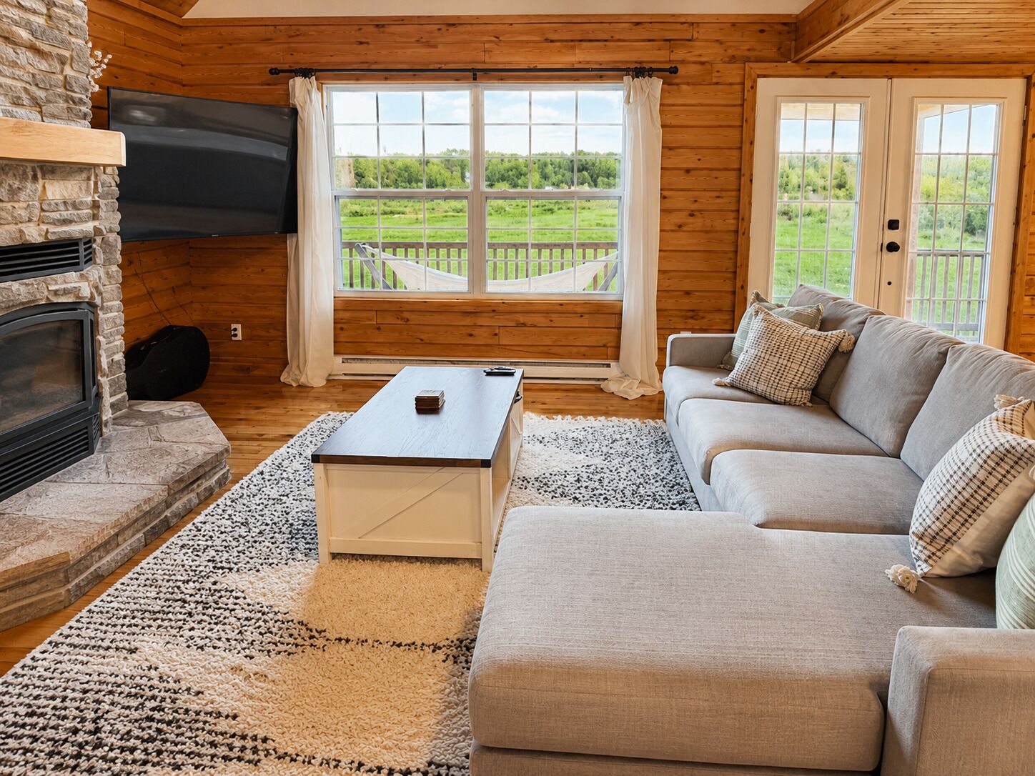 A log cabin interior with a stone fireplace, a flat-screen TV, a sofa, and a view of a green landscape through large windows.