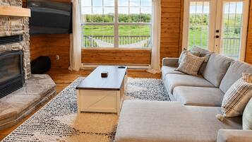 A log cabin interior with a stone fireplace, a flat-screen TV, a sofa, and a view of a green landscape through large windows.