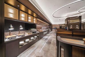 Een moderne hotellobby met een lange receptiebalie, een buffetruimte met diverse gerechten, en een zithoek met een hoge plafond en moderne verlichting.