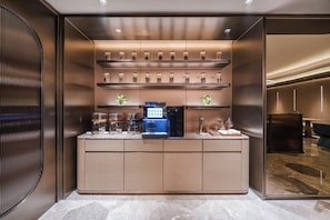 A modern, well-lit bar area with a coffee machine, shelves with jars, and a seating area with a sofa and table.