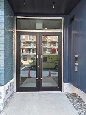 A building entrance with a glass door, a concrete ramp, and a visible street with a yellow vehicle and traffic cones. - The Panorama · Sleeps 9 · 2 baths · EV parking (Québec City)