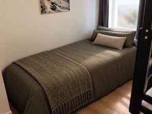 Ein Einzelbett mit einer strukturierten Decke und Kissen, ein Fenster mit Vorhängen und ein Holzboden.