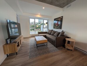 A modern living room with a sectional sofa, a flat-screen TV, a wooden coffee table, and a large window offering a city view. - The Panorama · Sleeps 9 · 2 baths · EV parking (Québec City)
