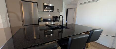 A modern kitchen with a central island, black bar stools, stainless steel appliances, and a visible air conditioning unit.
