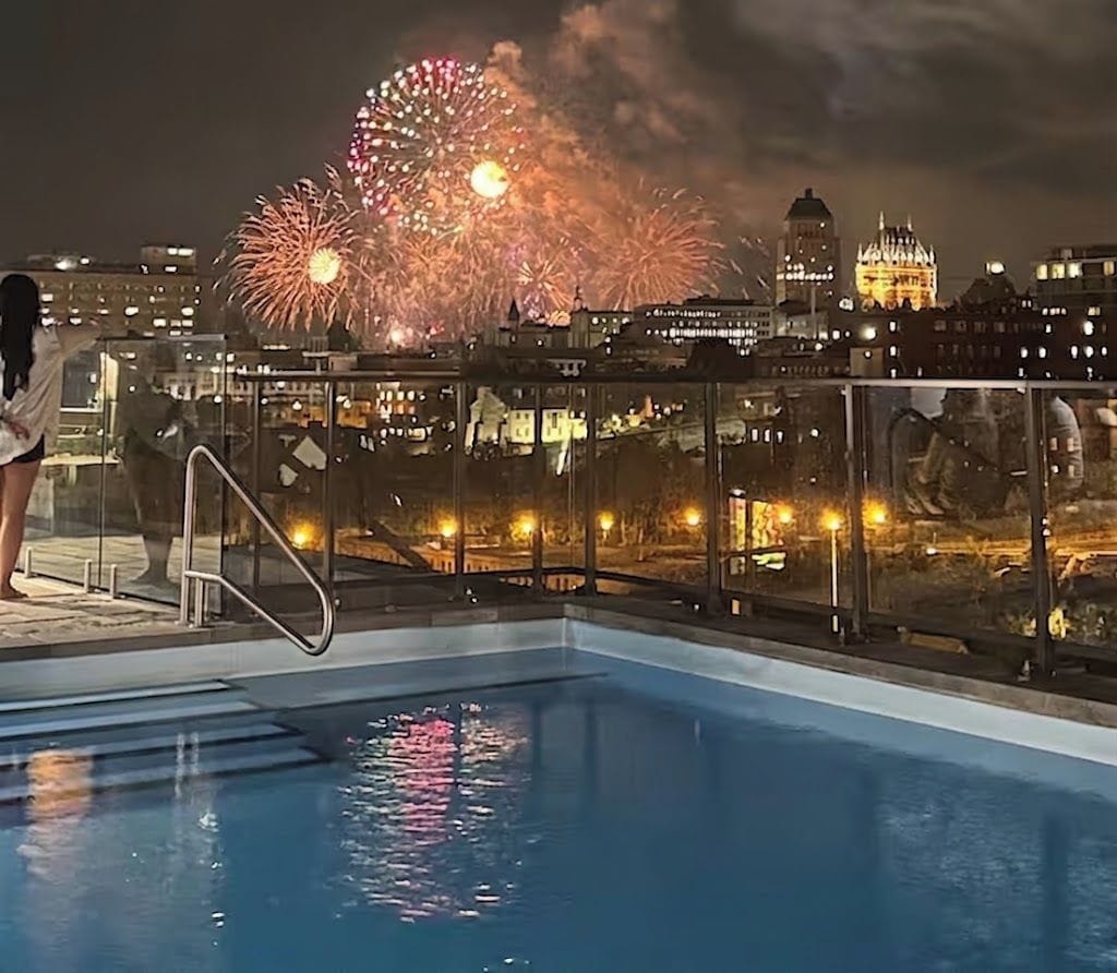 Une piscine dotée d’une balustrade en verre, un paysage urbain avec des bâtiments illuminés et des feux d’artifice en arrière-plan.
