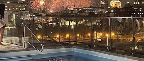 Una piscina con barandilla de vidrio, un paisaje urbano con edificios iluminados y fuegos artificiales al fondo.