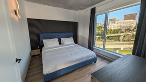 Een moderne slaapkamer met een groot bed, een nachtkastje, een bureau en uitzicht op een stadsgezicht door een groot raam.
