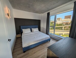 A modern bedroom with a large bed, a nightstand, a desk, and a view of a cityscape through a large window. - The Zenith · Sleeps 8 · 11th fl · EV parking (Québec City)