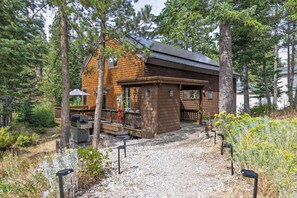 A cabin with a deck, surrounded by trees and gravel path.