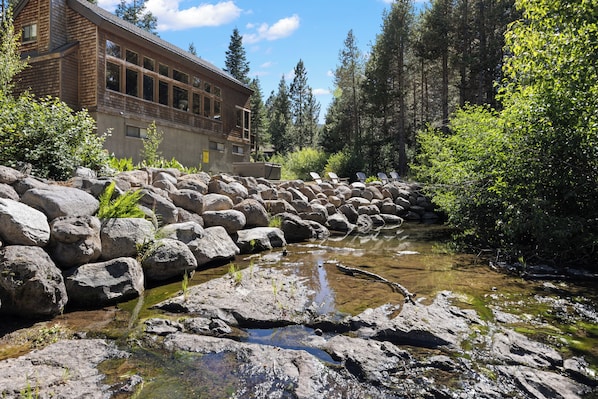 A natural landscape with a stream, rocks, and a building with large windows.