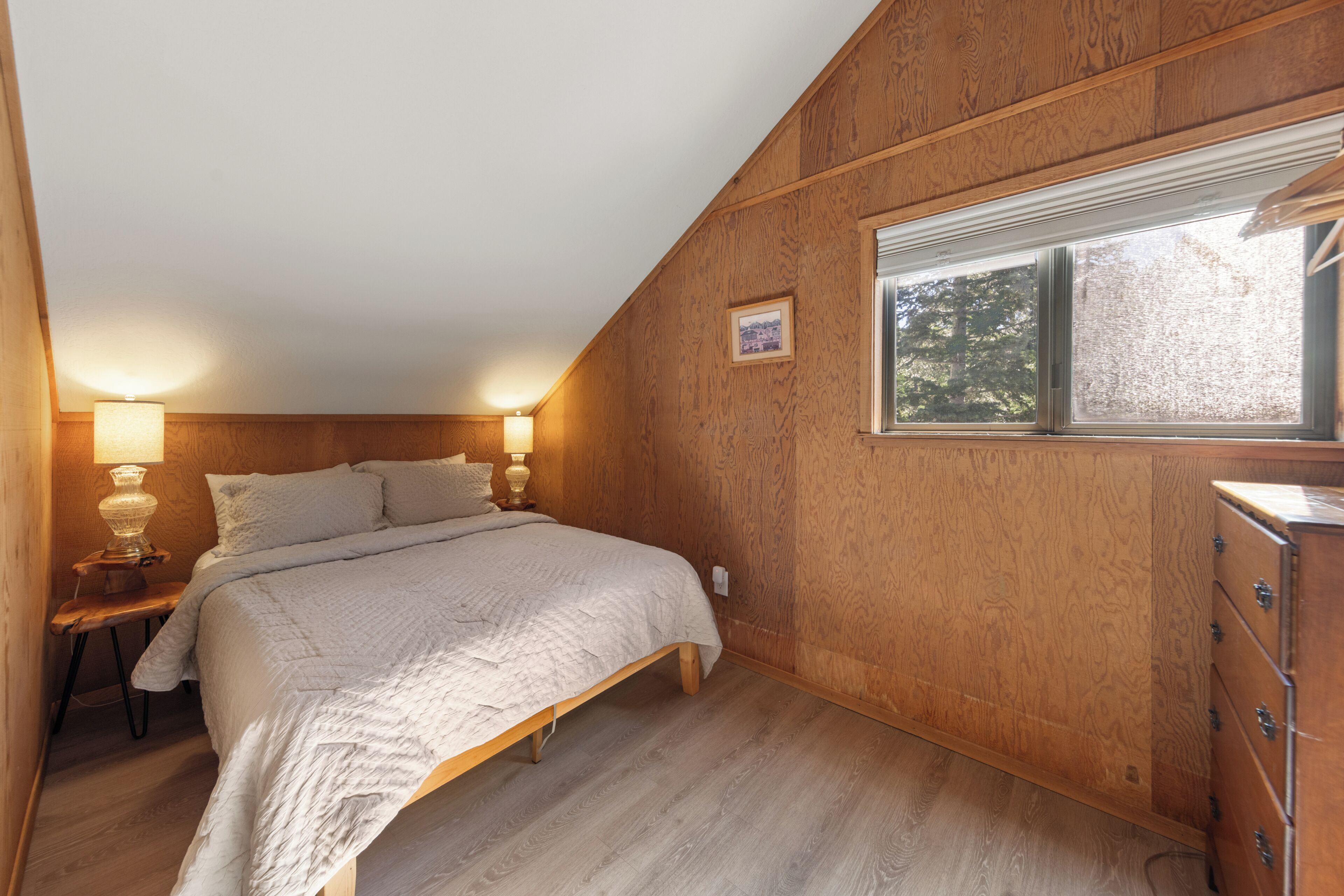 A bedroom with a bed, bedside lamps, a dresser, and a window with a view of trees.