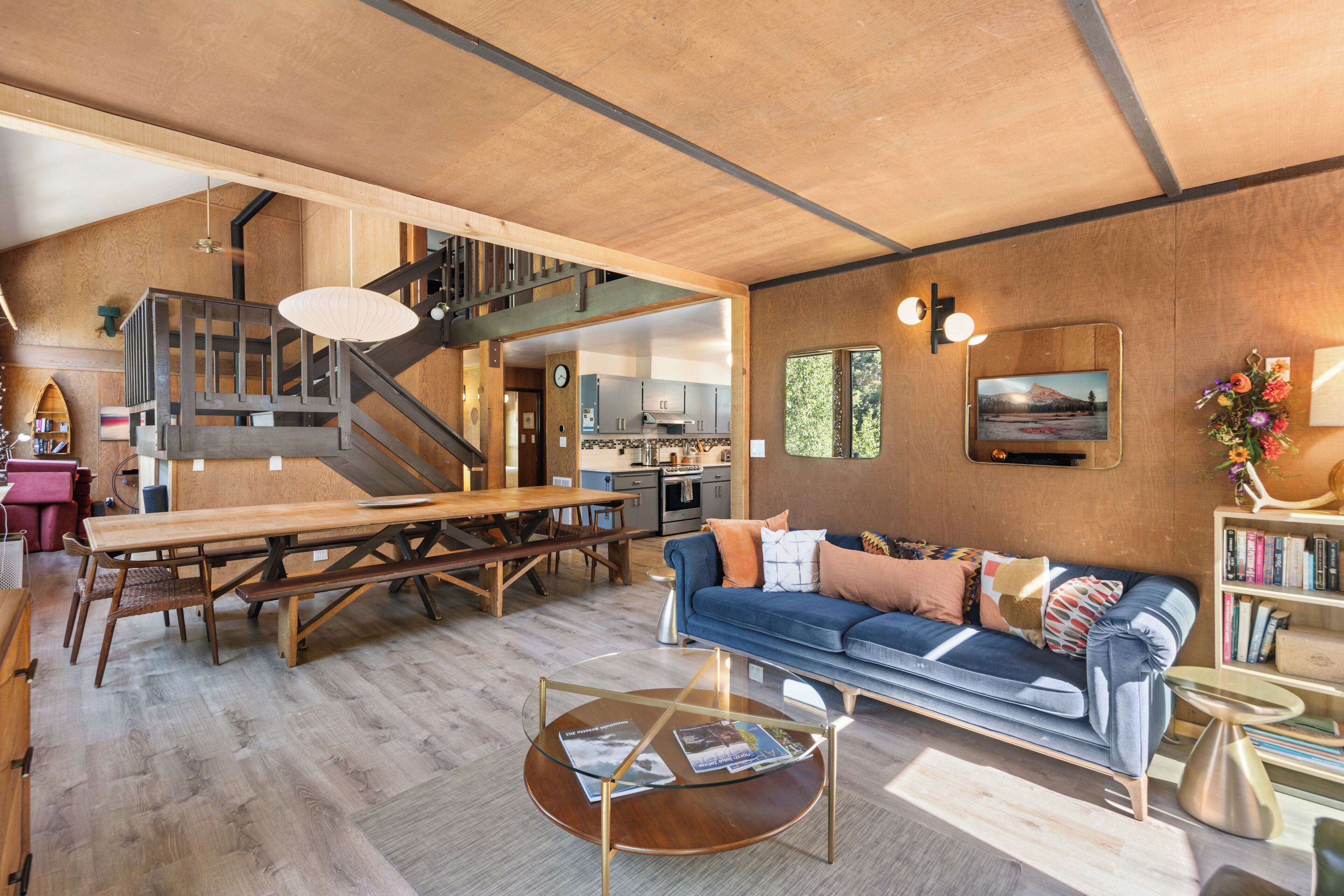 A spacious living area with a wooden staircase, a dining table, a sofa, and a kitchen in the background.