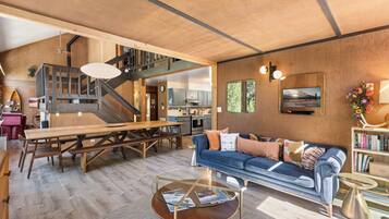 A spacious living area with a wooden staircase, a dining table, a sofa, and a kitchen in the background.