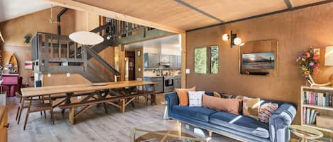 Un amplio salón con una escalera de madera, una mesa de comedor, un sofá y una cocina al fondo.