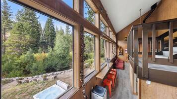A room with large windows overlooking a forest, a hot tub, and a seating area with red chairs.