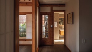 Un pasillo con vigas de madera, una puerta que da al baño y un ventanal con vistas a un muro de ladrillo y vegetación.