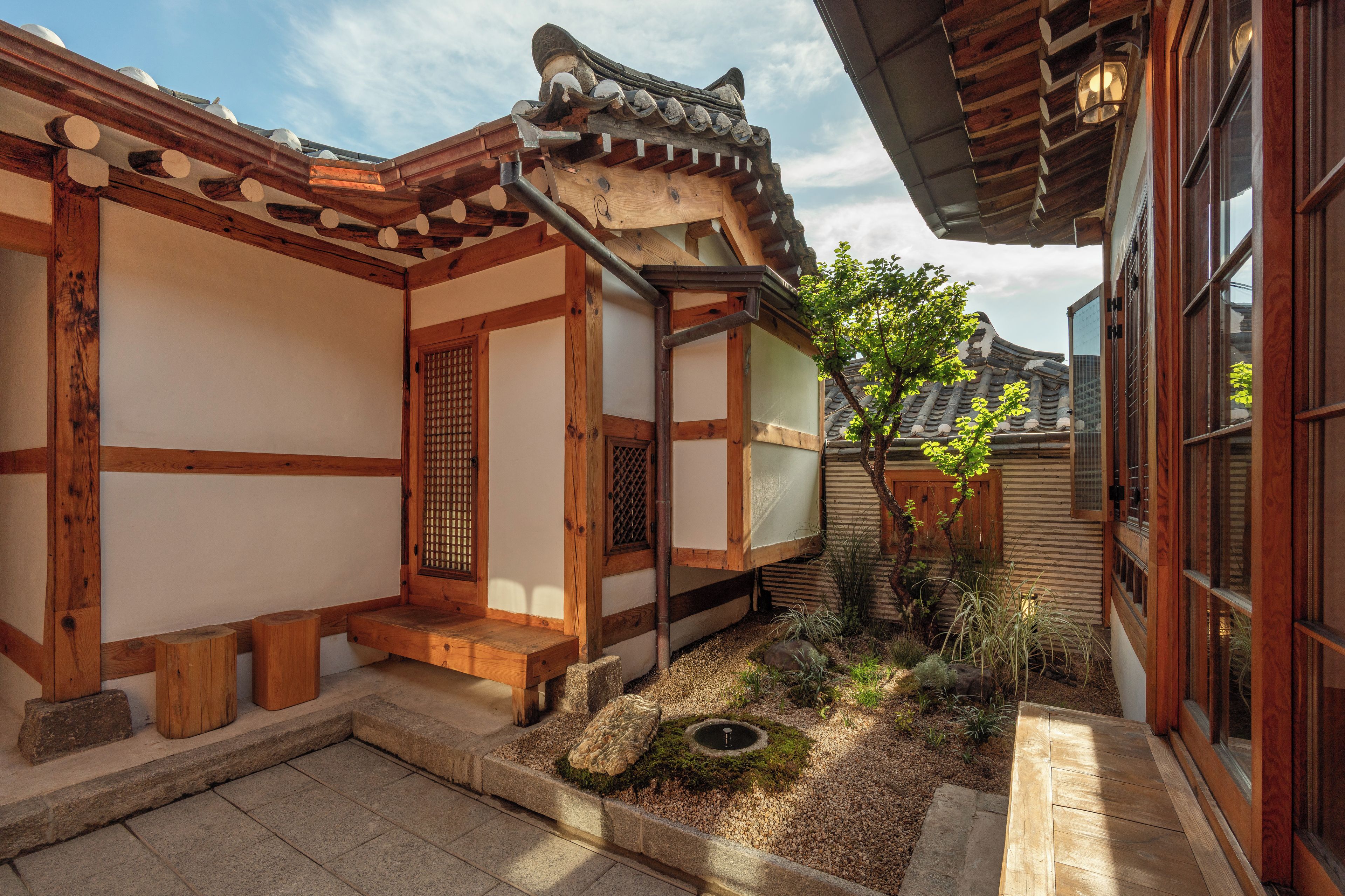A traditional Korean-style building with wooden beams, tiled roofs, and a small garden area with a tree and a stone lantern.