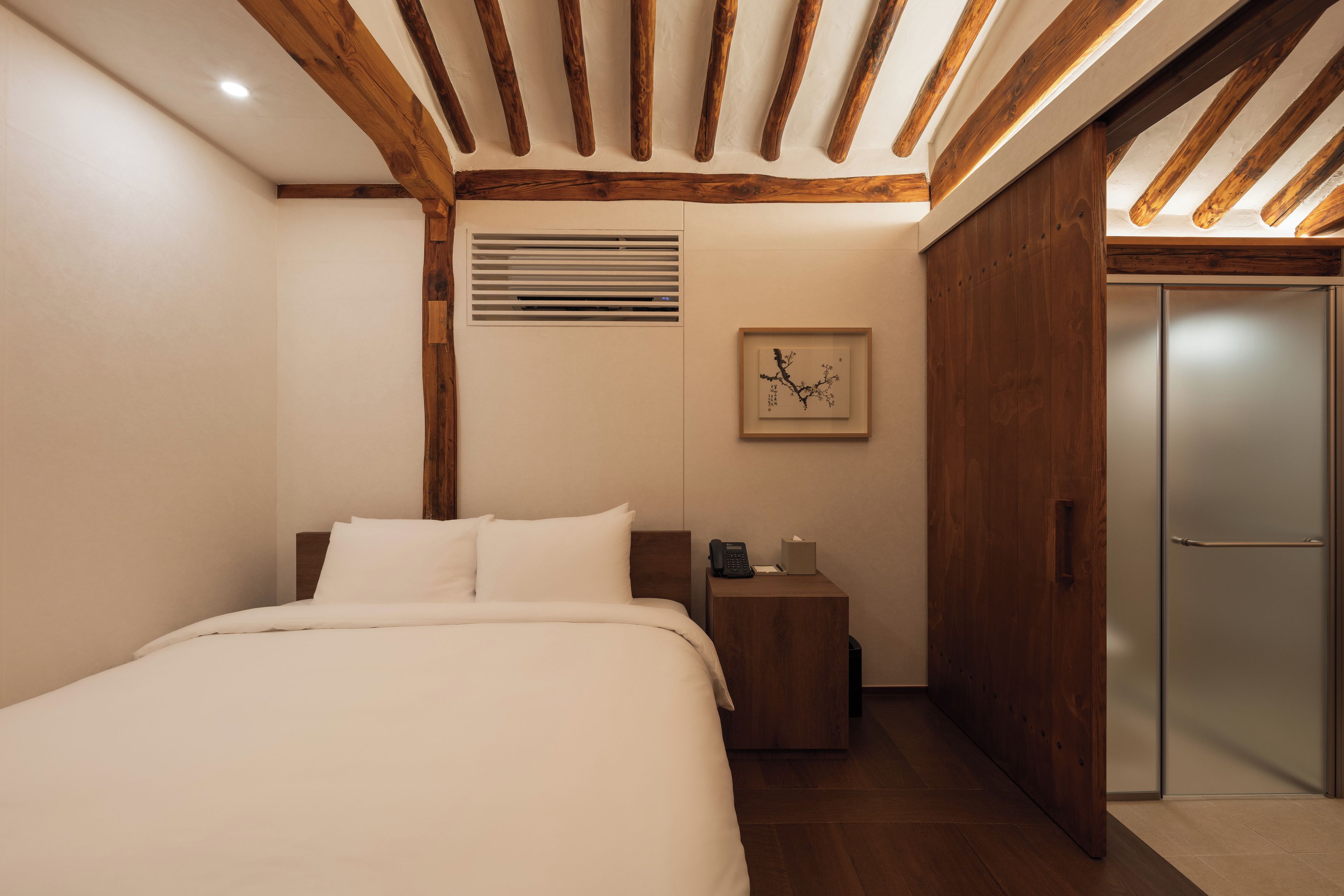 Un dormitorio con techo de madera, una cama con ropa de cama blanca, una mesita de noche con un teléfono y pañuelos, y una puerta.