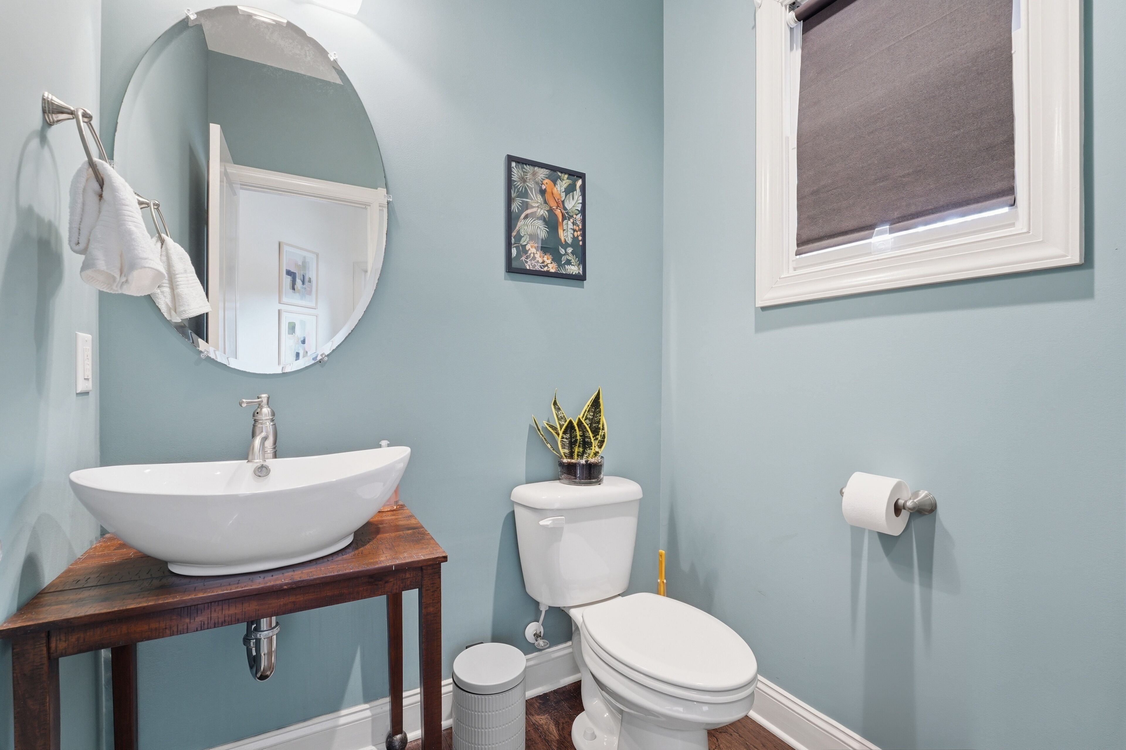 A bathroom with a white sink, a toilet, a small potted plant, a framed picture, and a window with a blind.
