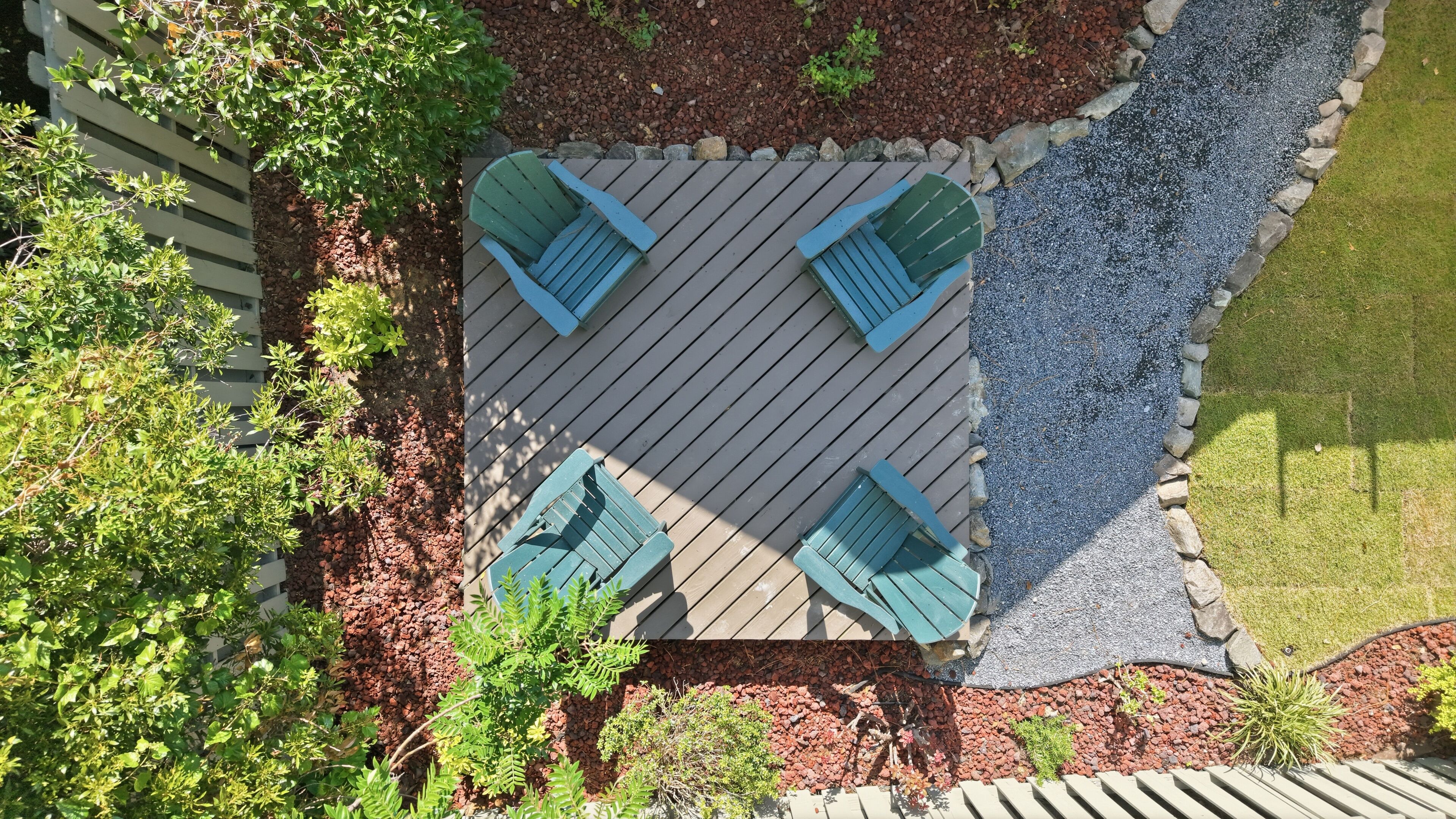 A top-down view of a patio area with a wooden deck, four green Adirondack chairs, and surrounding landscaping.