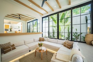 A modern living room with a large sectional sofa, a coffee table, and a view of lush greenery through large windows. - Villa Raya & Villa Viora Uluwatu (Pecatu)