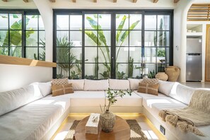 A modern living room with a sectional sofa, a coffee table, and a large window with tropical plants. - Villa Raya & Villa Viora Uluwatu (Pecatu)