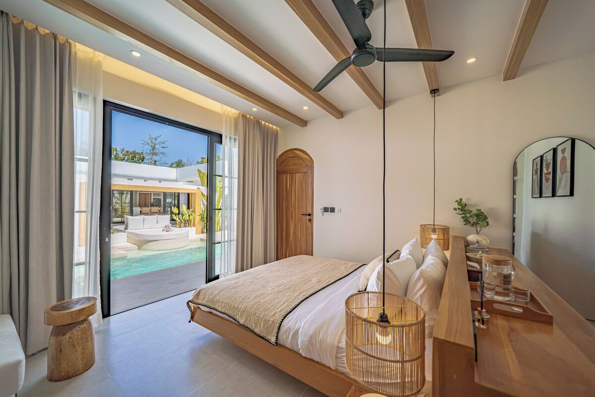 A modern bedroom with a large bed, a ceiling fan, a bedside table with a lamp, and a view of a pool and outdoor area.