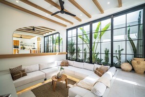 A modern living room with a large sectional sofa, a coffee table, and a view of tropical plants through large windows. - Villa Raya & Villa Viora Uluwatu (Pecatu)