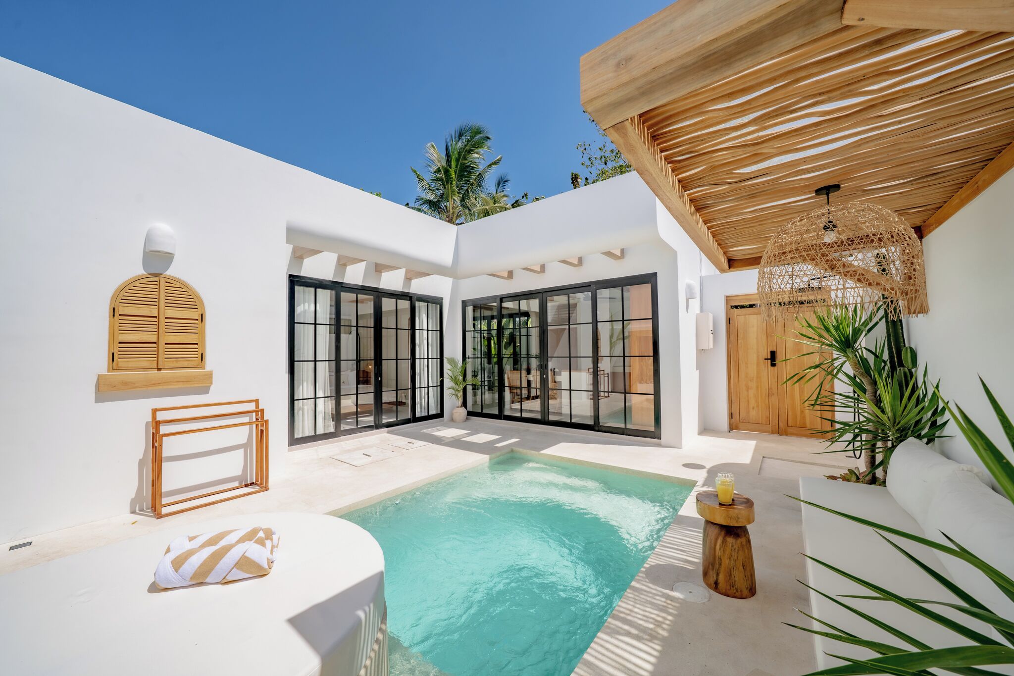 A pool area with a white wall, wooden pergola, and a wooden door.
