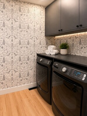 A laundry room with two front-loading washing machines, a wall with a patterned wallpaper, and a cabinet above the machines. - Cozy Stylish Home in Bridgeland (Calgary)