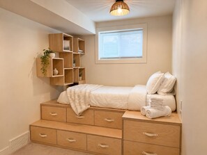 Ein Schlafzimmer mit einem Bett, einer Holzkommode mit Schubladen, einem Regal mit Pflanzen und einem Fenster mit Jalousien.