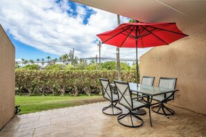 Eine Terrasse im Freien mit einem Glastisch und Stühlen, einem roten Sonnenschirm sowie Blick auf einen angelegten Garten und Palmen.