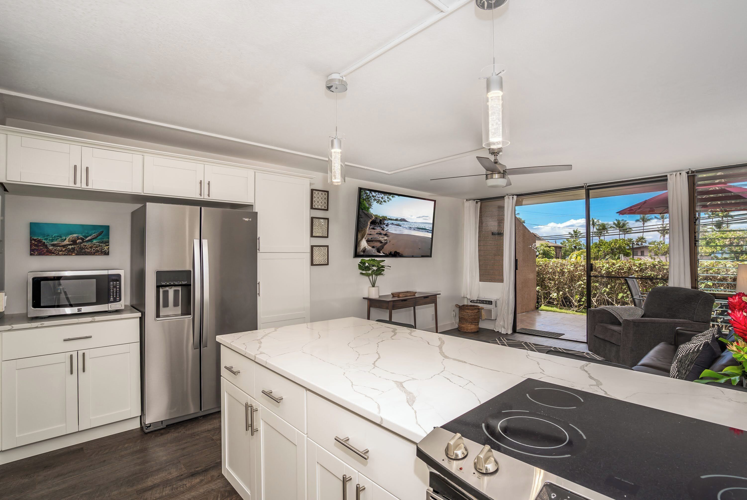A modern kitchen with white cabinetry, a large island with a black stovetop, a stainless steel refrigerator, and a microwave. There is a ceiling fan and a large window with a view of the outdoors.