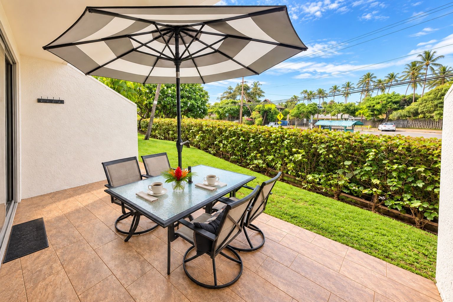 Une terrasse extérieure avec une table et des chaises, un grand parasol, et une vue sur un jardin bien entretenu et sur la rue.