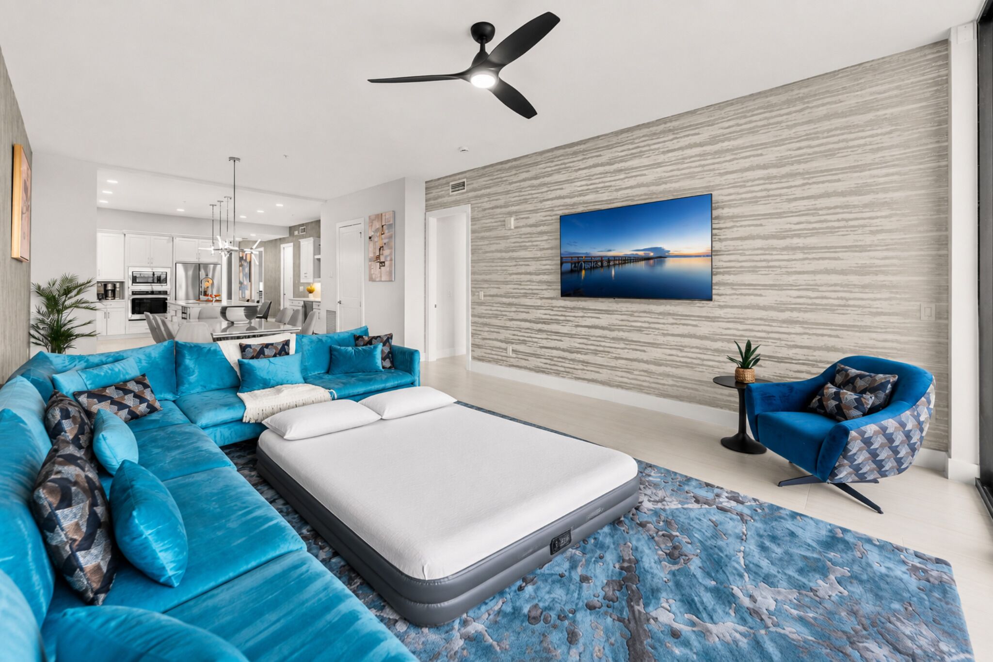 Een moderne woonkamer met een hoekbank, een enkel bed, een blauwe fauteuil en een groot schilderij aan de muur.
