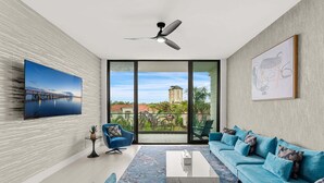 A modern living room with a large window, a ceiling fan, a blue sofa set, and a framed artwork. - Beautiful Melbourne Beach Retreat | Pool, Hot Tub & Private Balcony 🌴 (Melbourne Beach)