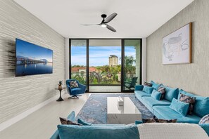 Ein modernes Wohnzimmer mit großem Fenster, Deckenventilator, blauem Sofagarnitur und einem gerahmten Kunstwerk.