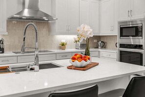 A modern kitchen with white cabinetry, stainless steel appliances, and a central island with a fruit bowl and flowers. - Beautiful Melbourne Beach Retreat | Pool, Hot Tub & Private Balcony 🌴 (Melbourne Beach)