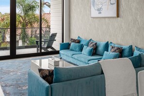 A modern living room with a blue sofa, patterned cushions, a glass coffee table, and a view of palm trees through large windows. - Beautiful Melbourne Beach Retreat | Pool, Hot Tub & Private Balcony 🌴 (Melbourne Beach)