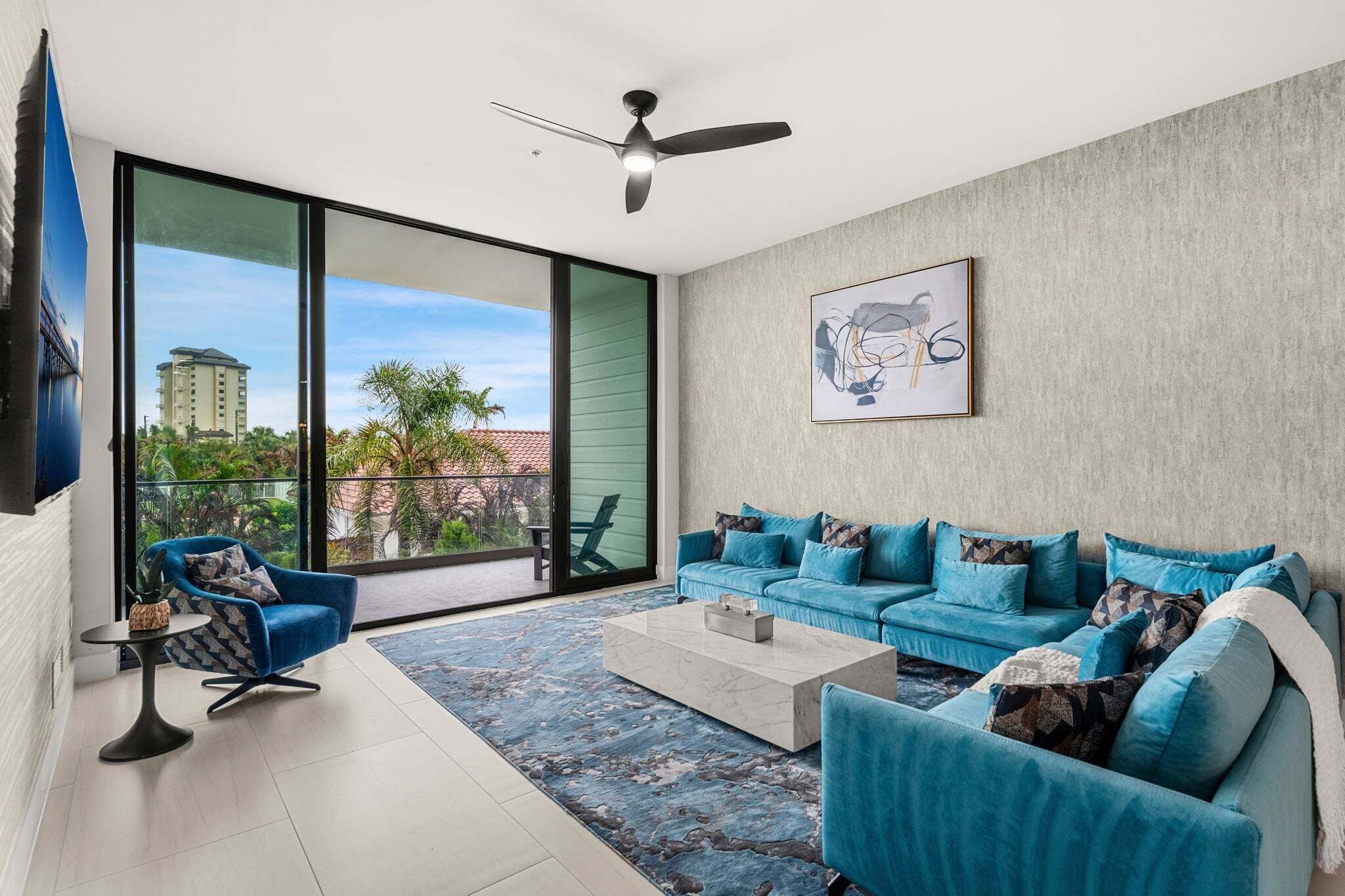 A modern living room with a blue sofa set, a ceiling fan, and a large window offering a view of buildings and greenery.