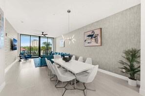 A modern dining area with a white table and chairs, a large window with a view of buildings and palm trees, and a ceiling fan. - Beautiful Melbourne Beach Retreat | Pool, Hot Tub & Private Balcony 🌴 (Melbourne Beach)