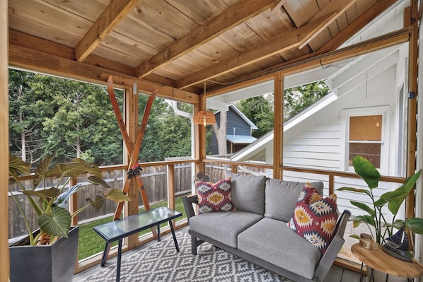 A covered porch with a gray sofa, patterned cushions, a glass table, and potted plants.