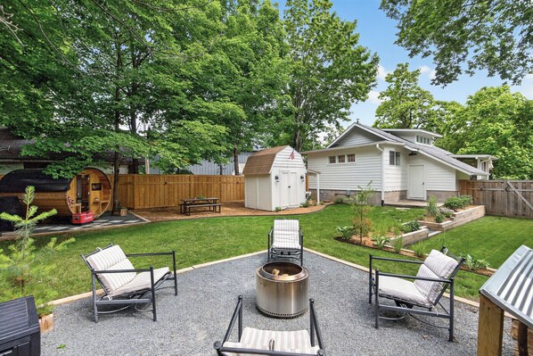 A backyard with a fire pit, two lounge chairs, a wooden shed, and a house.